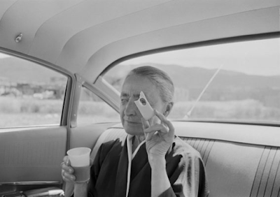 black and white photograph of Georgia O'Keefe inside a car holding a piece of Swiss cheese up to her eye