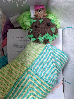 in-progress mitered-square blanket knit in pastel colors sitting atop the pattern and other yarn.  There is also a small knit groundhog poking out the top of its knit mound and hole