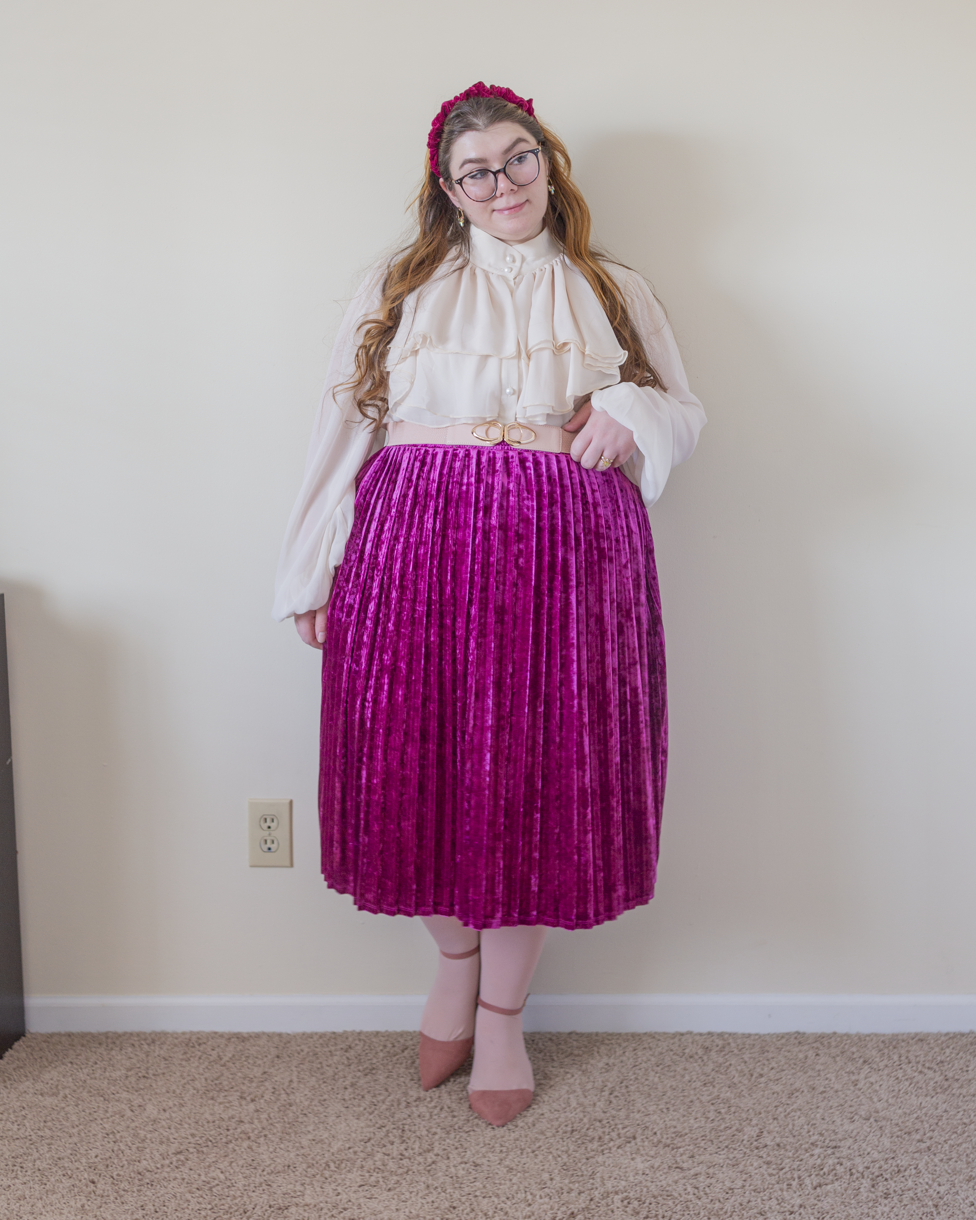 An outfit consisting of a cream sheer bishop sleeve blouse with a neck ruffle, tucked into a berry pink velvet pleated midi skirt and pink ankle strap heels.