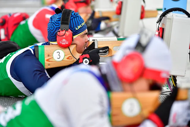 Atleta deficiente visual ucraniano atira deitado usando um rifle. Ele usa um gorro azul e amarelo escrito Ucrânia em inglês e um par de fones de ouvido vermelho, que ele utiliza para guiar a sua mira