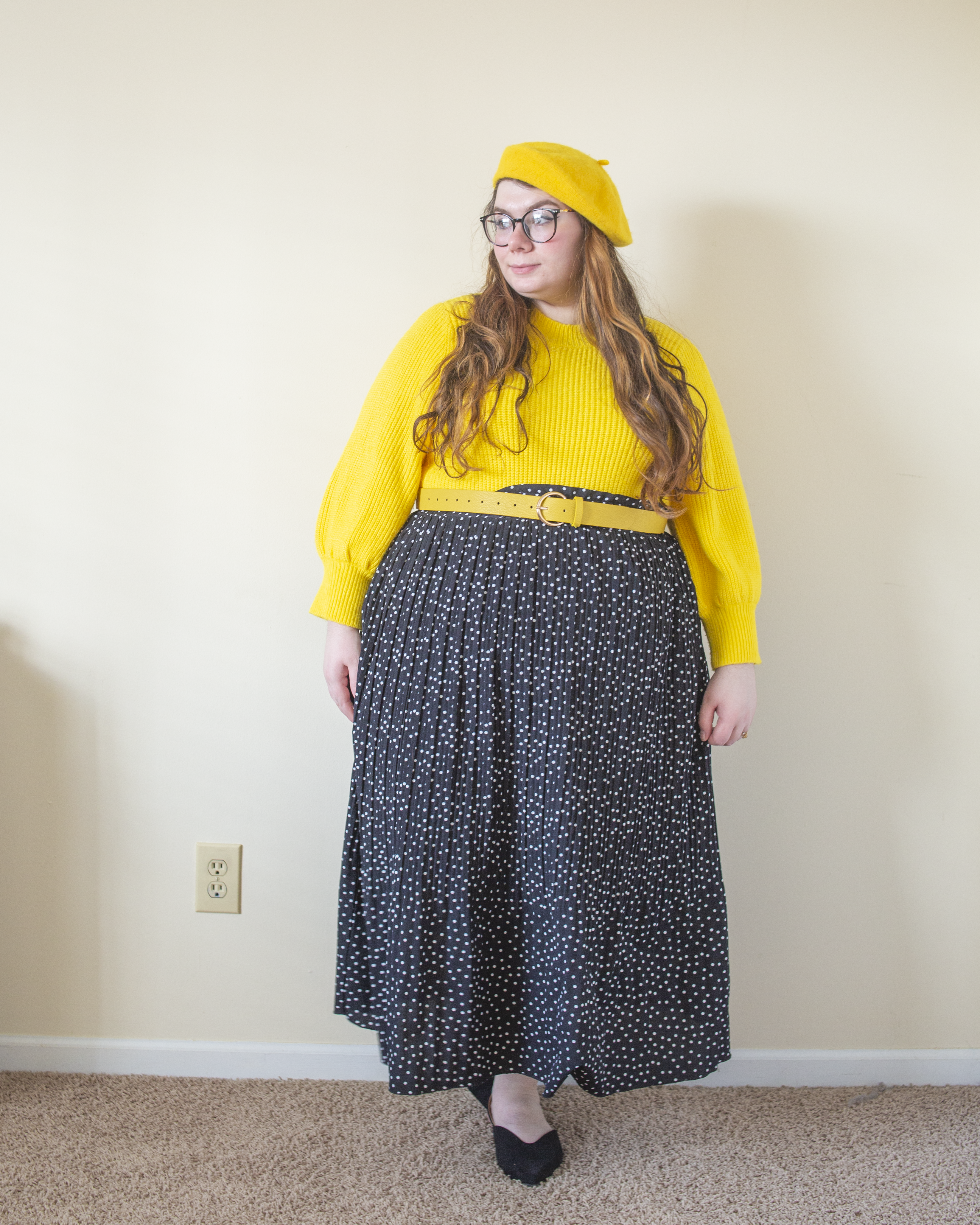 An outfit consisting of a yellow wool beret, a black leather jacket over a yellow high neck balloon sleeve sweater tucked into a white on black micro swiss dot midi skirt.