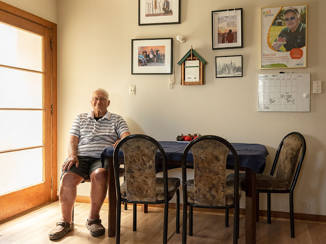 Man sitting at a table in his house. There are pictures on the wall above him.