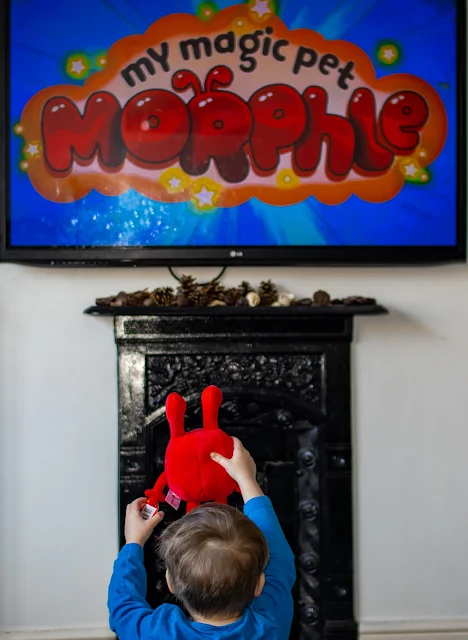 A toddler sitting in front of the television with a morphle plush toy watching Morphle