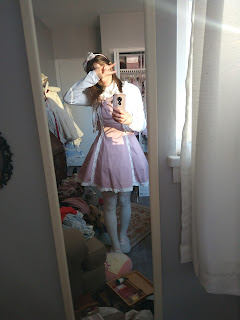 The blogger is standing in front of a mirror taking a photo in a lolita dress with a cardigan. They are covering their face with a peace sign. They're standing in a messy room.