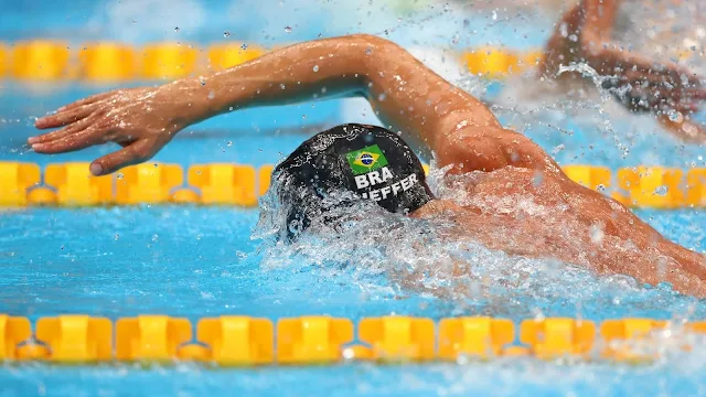 Fernando Scheffer durante prova de natação. Apenas metade da cabeça e um braço do nadador estão fora da água. Ele usa uma touca preta com a bandeira do Brasil e seu sobrenome