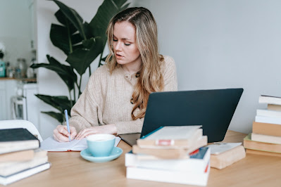 Girl with laptop at desk surrounded by books writing in notepad. Photo by George Milton from Pexels