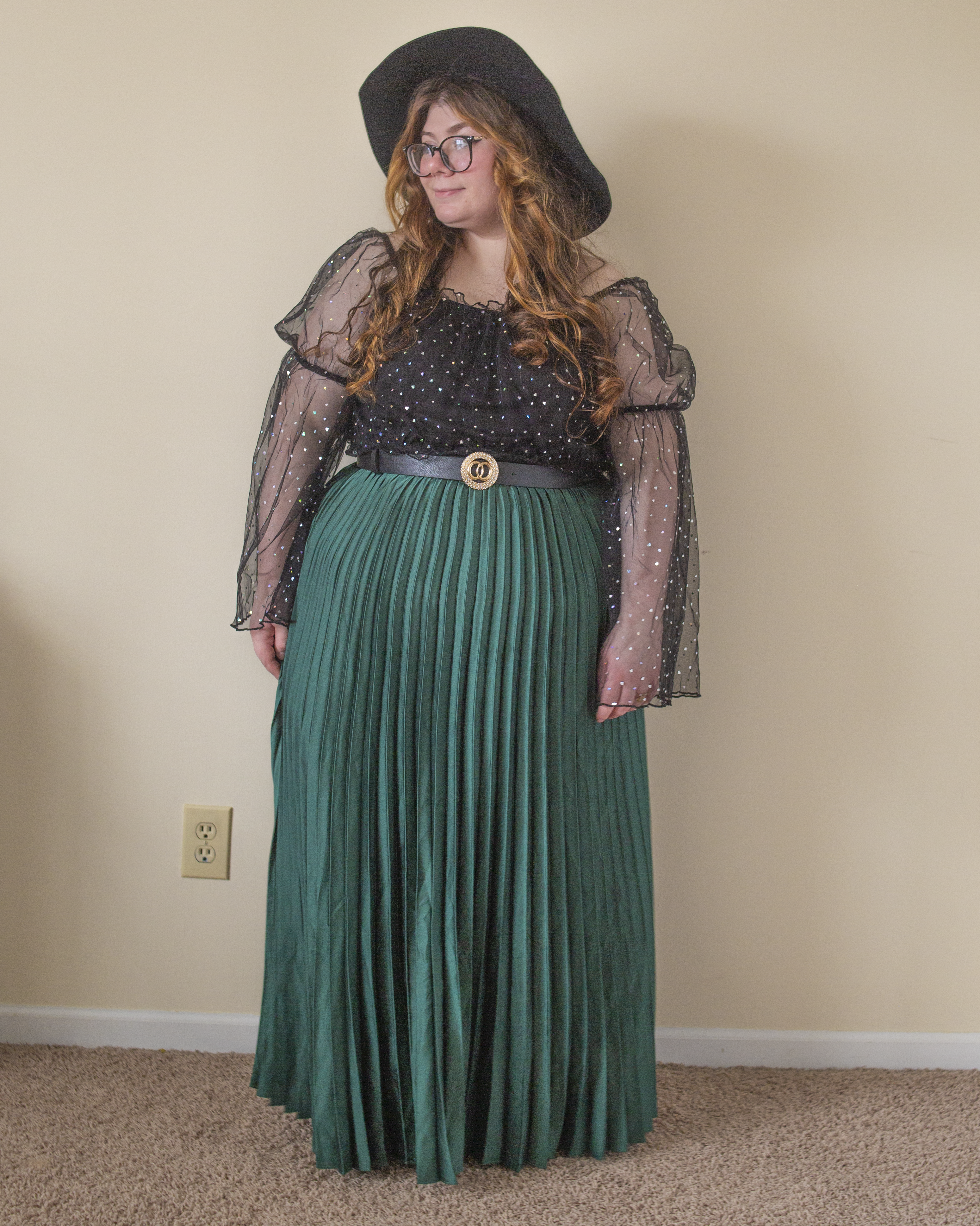 An outfit consisting of a black sheer blouse with bell sleeves and silver holographic heart print, tucked into a green pleated maxi skirt and black ankle boots.