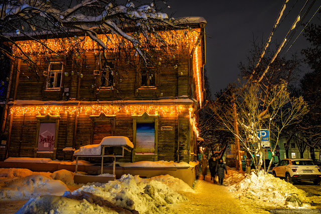 Пешеходы идут по дорожке вдоль деревянного дома с новогодней подсветке