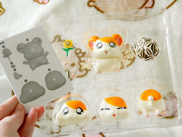 A photo showing a nendoroid figure in its plastic packaging. The character is an orange and white hamster, and there are various head and body parts to assemble. The blog writers hand can be seen holding a small instruction manual