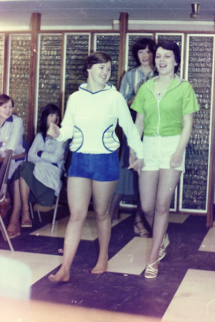 One staff member dressed in a white and blue hooded top and blue shorts, followed by another wearing a green zip up short-sleeved shirt and white high rise shorts