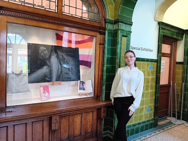 Fern stood next to their finished exhibition, which contains their artwork, chest binder, Grayson's Art Club exhibition catalogue and their crocheted Lesbian Pride flag
