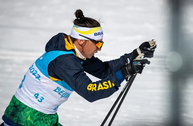 Wesley, usando um coque no cabelo, esquia com um fundo branco de neve. O brasileiro veste um agasalho do CPB com azul com detalhes em verde e amarelo, com um colete com o número 43 por cima.