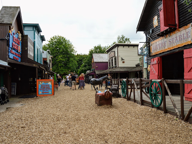 Image of The Wild West area of Sundown Adventureland. The image shows a bark paved street lined with buildings that look like they are from the wild west. They look as though they are made of wood and they house attractions such as play areas and a soft play. There are model horses, cowboys and bandits dotted along the street as well as a drunk character asleep in a drinking trough.