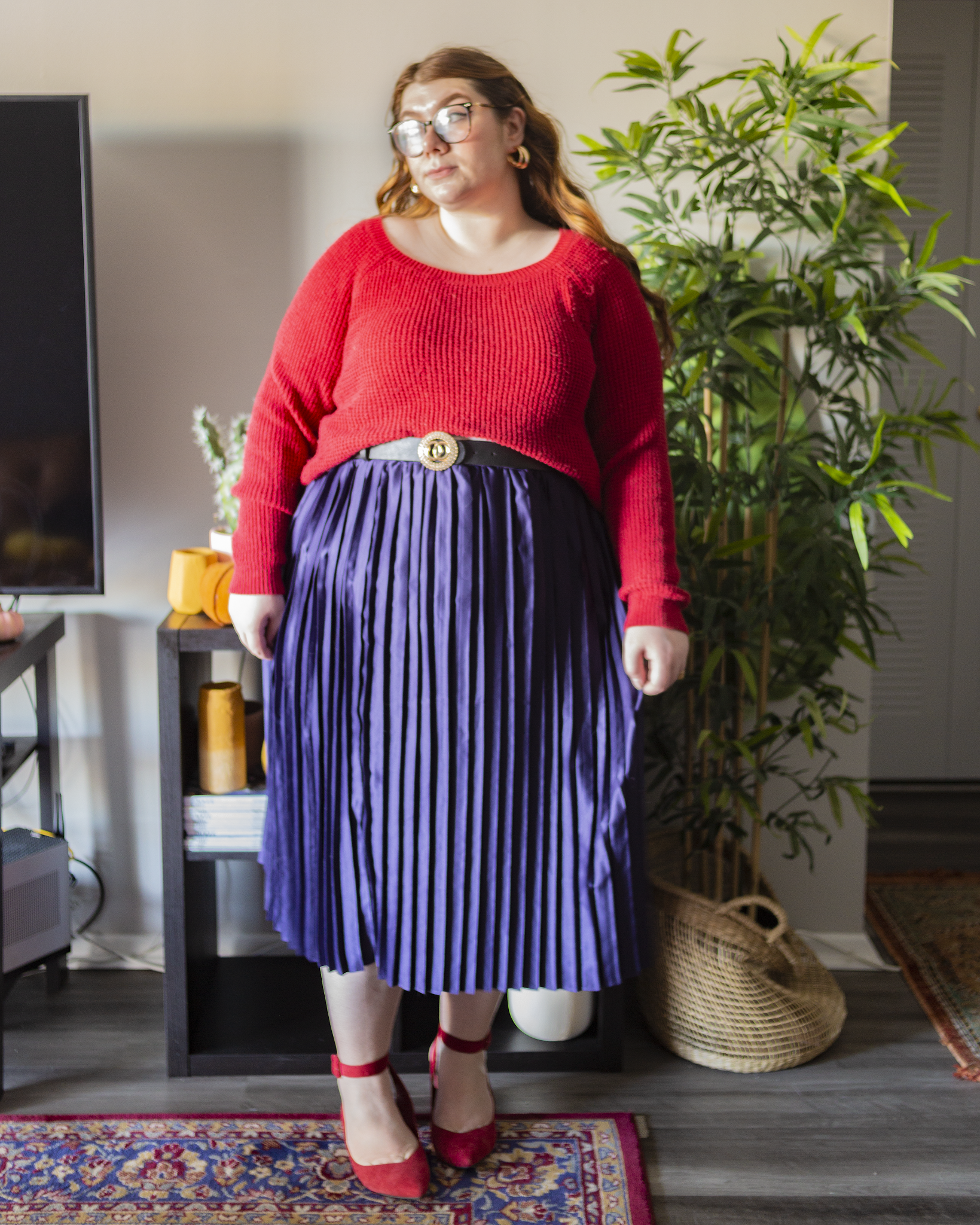 An outfit consisting of a red knit sweater half tucked into a navy blue satin pleated midi skirt and red heels.