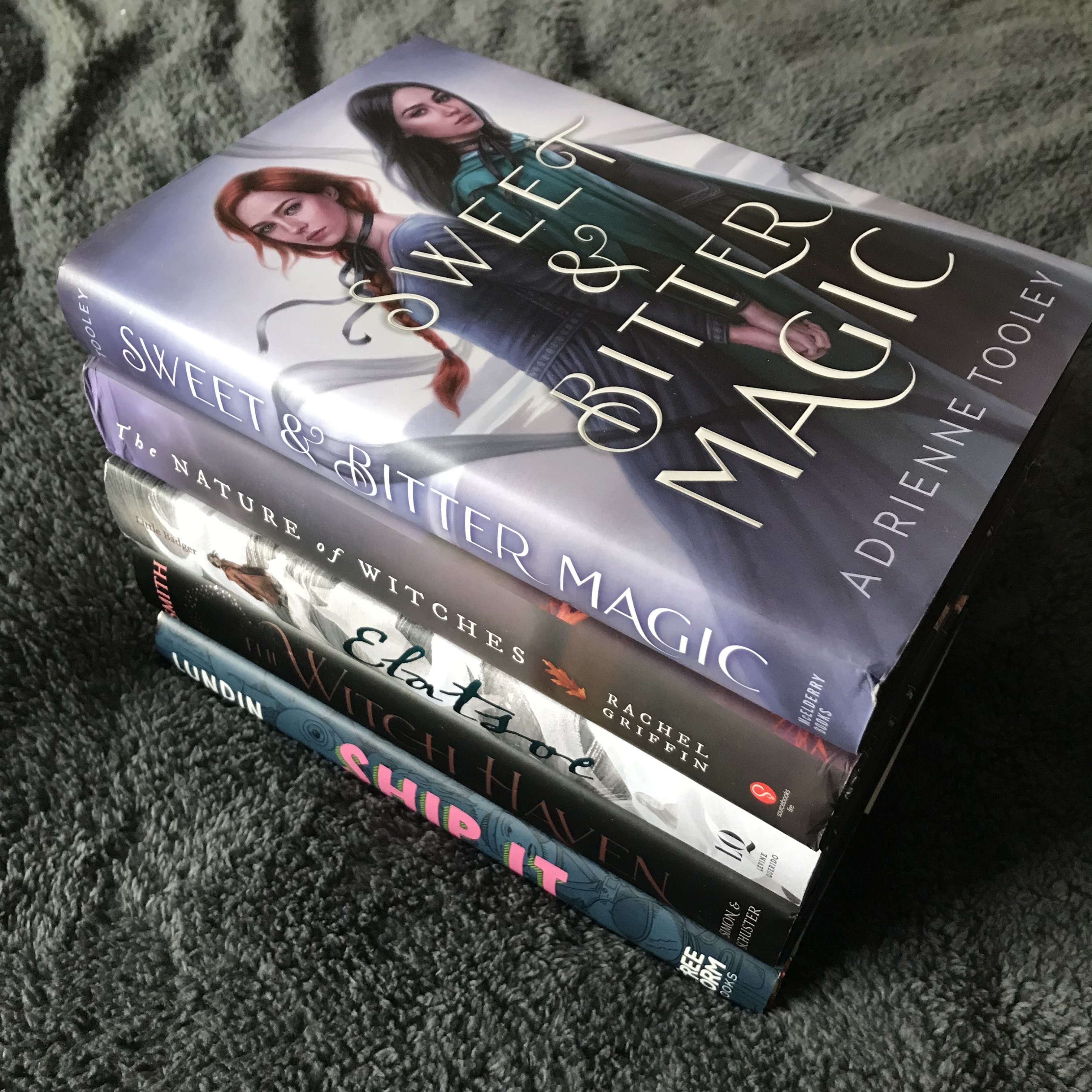 A stack of hardback books on a fluffy, dark grey duvet cover. The books from top to bottom are Sweet & Bitter Magic by Adrienne Tooley, The Nature of Witches by Rachel Griffin, Elatsoe by Darcie Little Badger, The Witch Haven by Sasha Peyton Smith, and Ship It by Britta Lundin. The photo is taken at an angle from above, so the spines are on show. The spines face the bottom left corner of the photo.