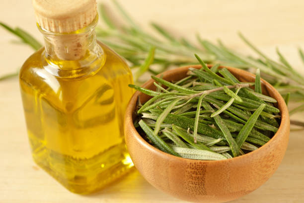 A petite bottle of fragrant rosemary-infused oil accompanied by fresh rosemary leaves displayed in a rustic wooden bowl.