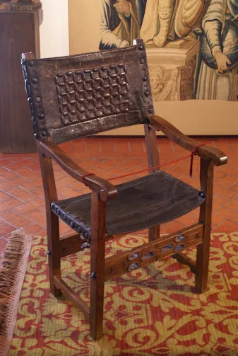 Fotografía del auténtico Sillón del Diablo, un sillón de madera oscuro de estilo frailero, con respaldo y asiento de cuero decorado con tachuelas y grabados. El sillón está exhibido sobre una alfombra de motivos rojos y amarillos, con una cuerda roja que va de un reposabrazos al otro, restringiendo el acceso. Detrás se distingue un tapiz antiguo que complementa el ambiente histórico y misterioso de la escena.