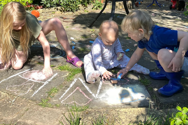 3 children on various ages from baby upwards drawing with chunky pastels on paving in a garden