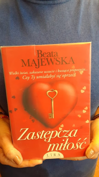 Pani w szarej bluzce prezentuje książkę Beaty Majewskiej pod tytułem Zastępcza miłość.