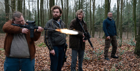 Still from Riders of Justice: the four actors left-to-right: Nicolas Bro, Nikolaj Lie, Lars Brygmann and Mads Mikkelsen stand in the woods. Mads looks on as the other three men hold assault rifles (this is a practice range) They're all looking at Nicolas Bro who is firing his gun.