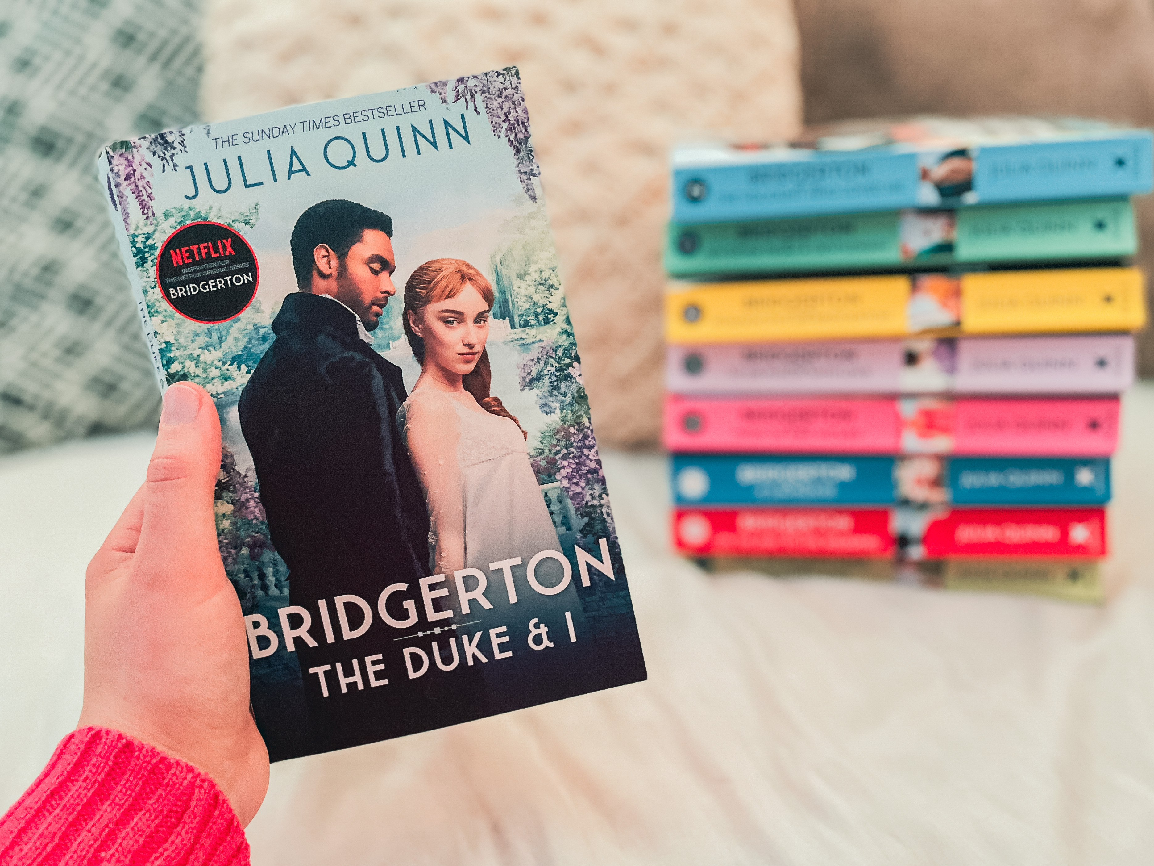 Hand holding Bridgerton: The Duke and I by Julia Quinn in front of stack of rest of Bridgerton book series