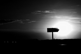 A sign pointing at the sun during a sunset.