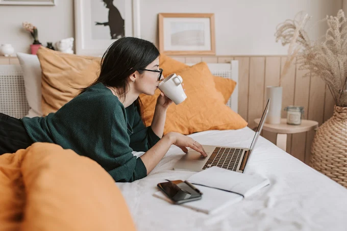 Mulher deitada na cama, tomando café e usando laptop