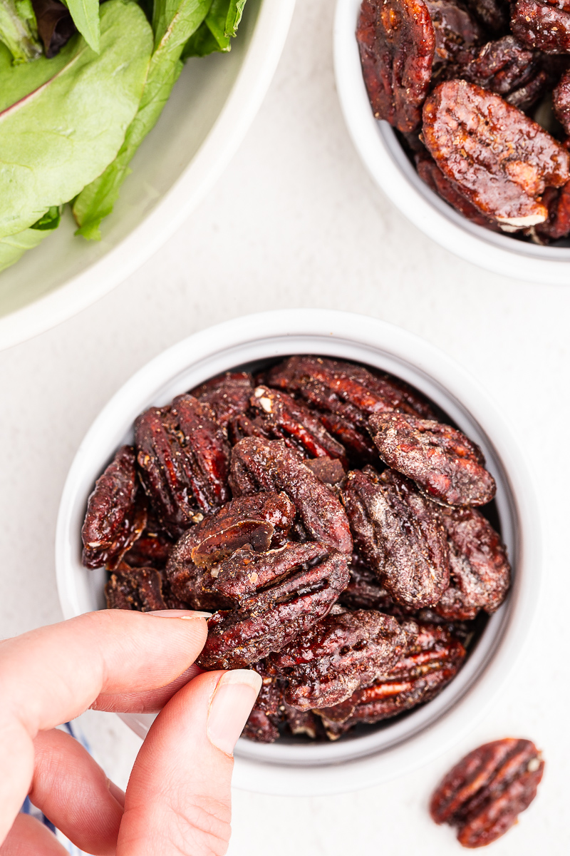 Photo of a hand holding a keto candied pecan above a white bowl filled with the candied pecans.