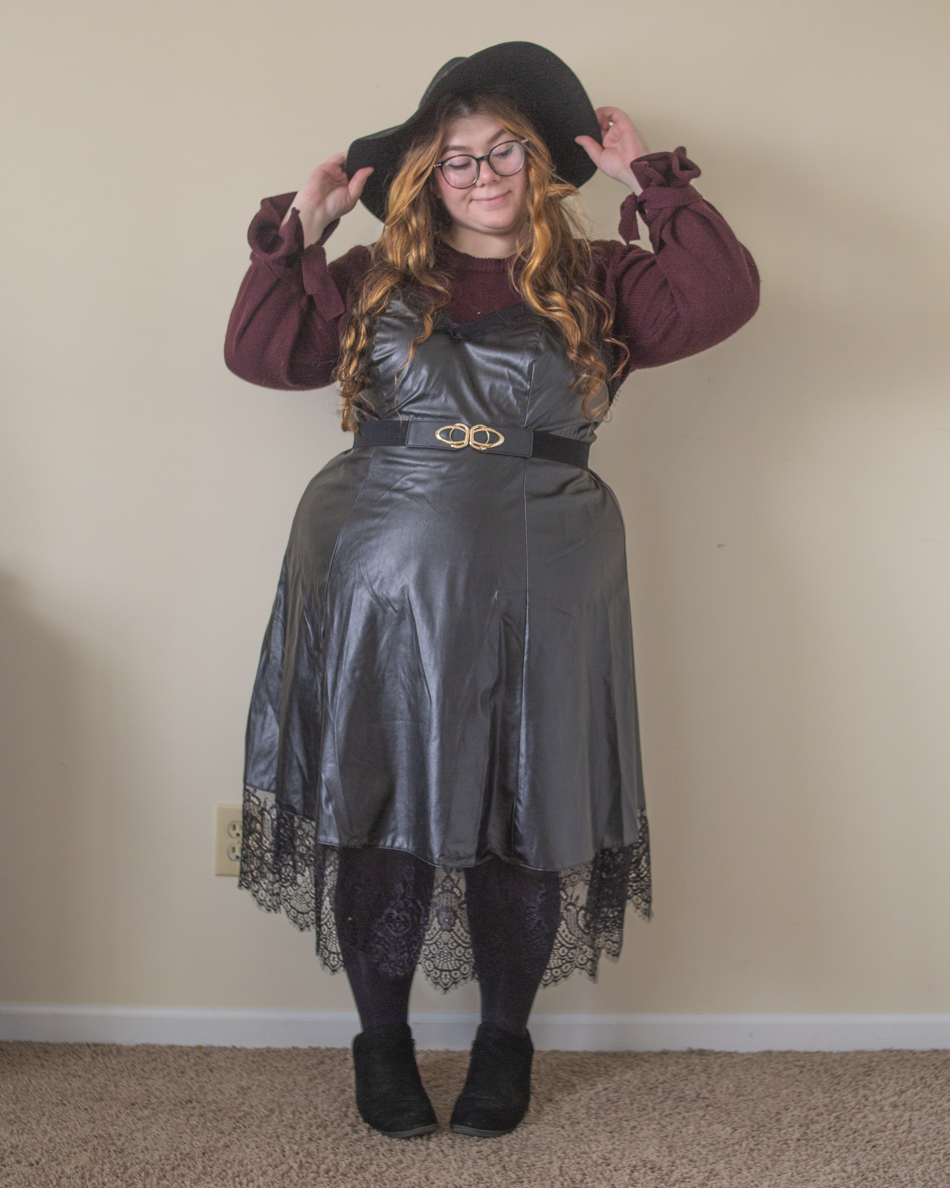 An outfit consisting of a berry knit sweater with ties around the wrists under a black leather midi dress with black lace trim, black tights and black boots.