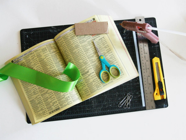 supplies for making a phone book letter holder including cutting mat, phone book, wide ribbon, scissors, glue gun, paper clips, metal ruler, craft knife
