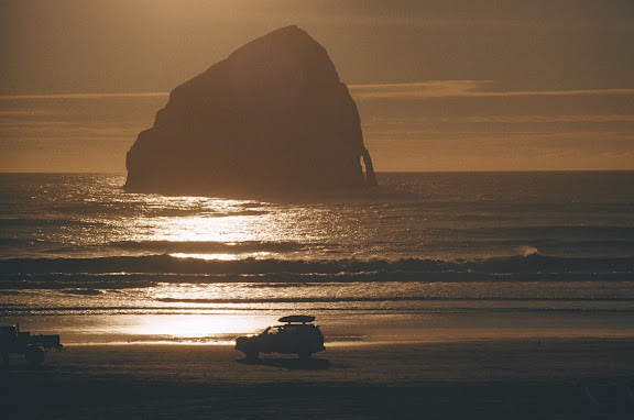 Ocean beach scene, with sun setting beyond huge rock rising from the sea, and a car with surfboards on the roof driving along the water's edge.