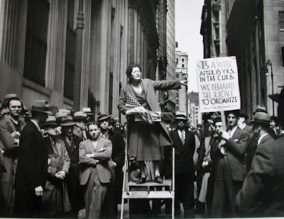 black and white photograph of a young woman Union organizer on a step stool giving a speech to office workers on the lunch break in New York's Wall Street area, 1936