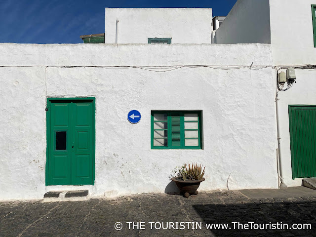 A street sign of an arrow that points to the left, between a green wooden door and a window in the same colour on a whitewashed flat-roofed cottage under a blue sky.