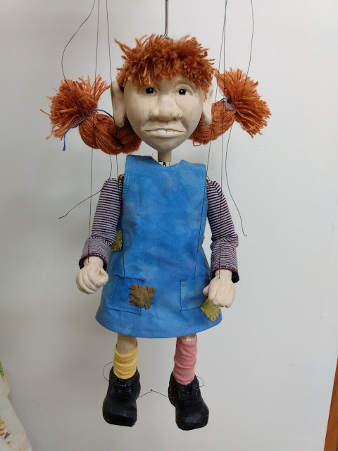 A simple marionette styled like Pippi, with thick red braids, a blue sleeveless dress over a striped long-sleeved t-shirt.