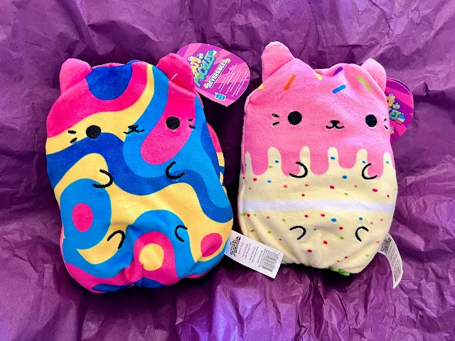 a cat plush with pink, yellow and blue swirls and a cat plush that looks like a cake with pink icing and sprinkles