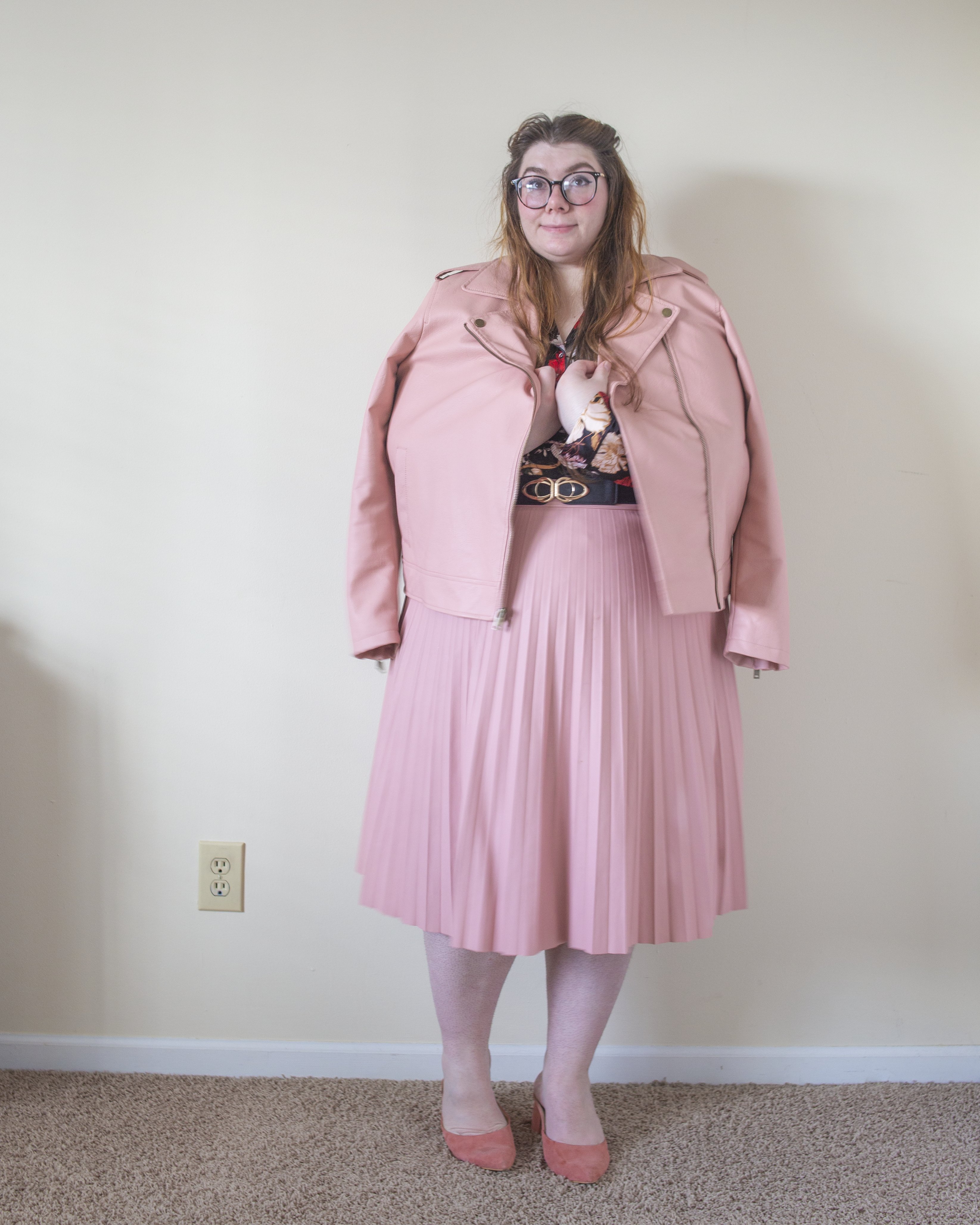 An outfit consisting of a pastel pink faux leather jacket over a black, cream, pink, yellow and red pajama style satin top tucked into a pastel pink leather pleated midi skirt and pink heeled clogs.