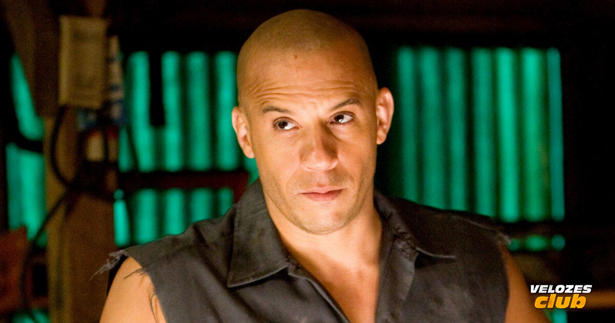 Na imagem vemos o ator Vin Diesel com uma camisa preta sem manga olhando para o lado em uma garagem.