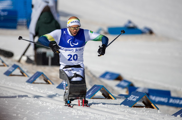 Robelson esquia usando o esqui adaptado. Ele é um homem careca, vestindo uma malha azul e branca e um colete com o número 20. Ele também usa uma faixa na testa escrito  Brasil