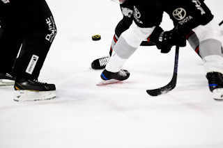 Ice hockey being played. One player is holding a hockey stick. The puck is in the air, and you can only see the players heads.