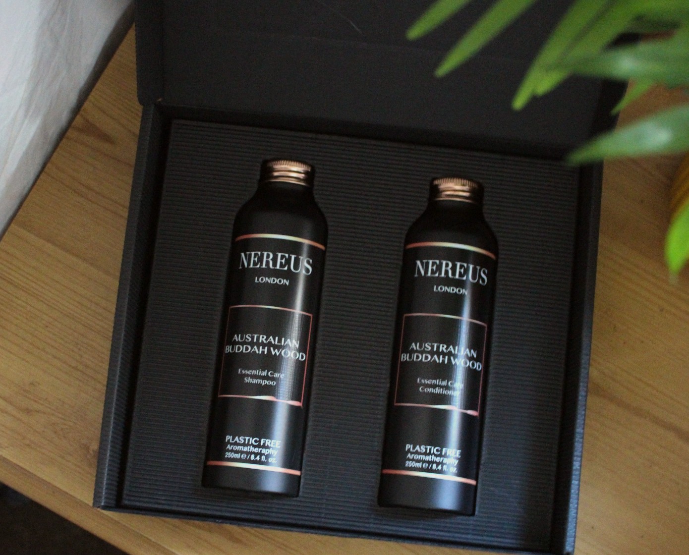 The image shows a black box on a brown wooden surface. The black box contains two black metal bottles with rose gold lids. One bottle reads "Nereus London Australian Buddhawood Essential Care Shampoo" whilst the other read "Nereus London Australian Buddhawood Essential Care Conditioner".