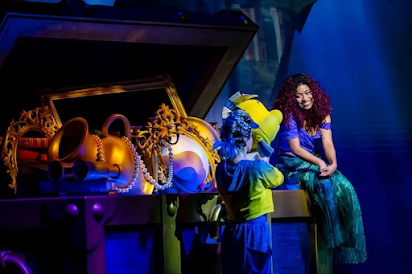 An Ariel performer sits next to a little boy holding up a Flounder puppet on a giant treasure chest.