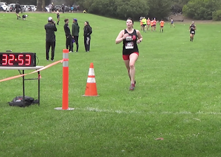 Me running toward the finish line of a cross country race in red shorts, a black tank, and race bib against a background of green grass and other runners in the distance.