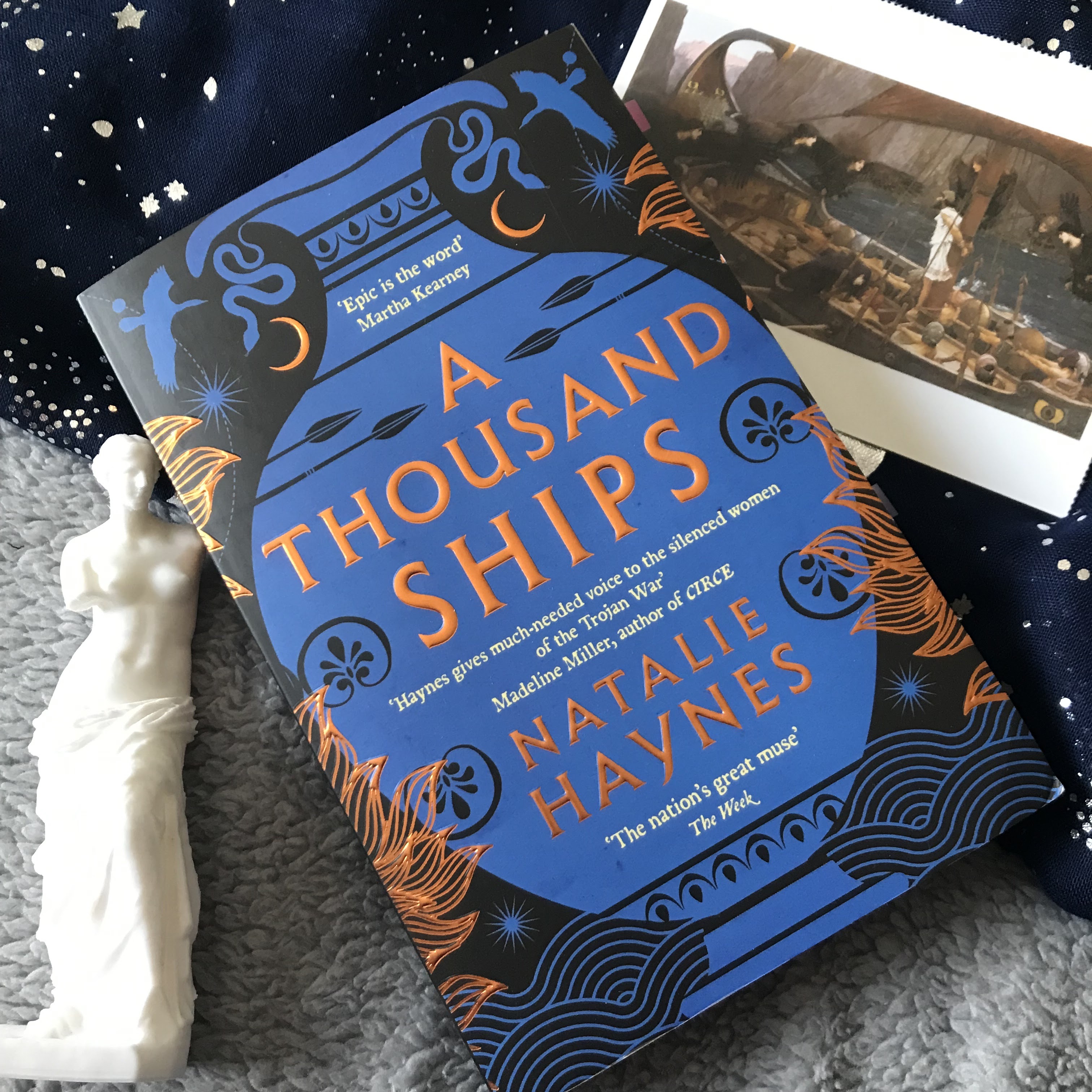 A photo of A Thousand Ships by Natalie Haynes, diagonal, with the top facing the top left corner and the bottom facing the bottom right. It's half on a dark blue scarf with metallic silver stars and suns, and half on a fluffy, light grey duvet. On it's right, and slightly underneath, is a postcard of Ulysses and the Sirens by John William Waterhouse, and on it's left is a white Venus de Milo ornament.