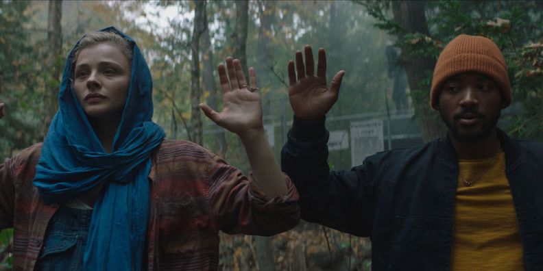 Dois personagens do Filme Mãe x Androides estão presentes na imagem, estão se rendendo com as mãos levantadas, apresentam uma expressão de medo