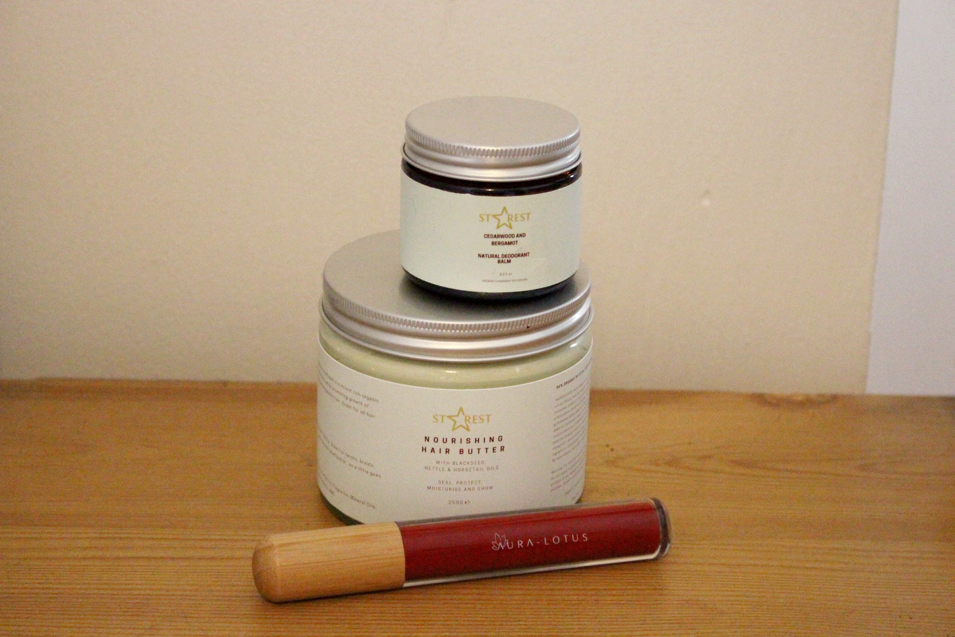 The photo shows a small jar of deodorant balm stacked on top of a large jar of hair butter. In front is a red lipstick with a brown wooden lid.