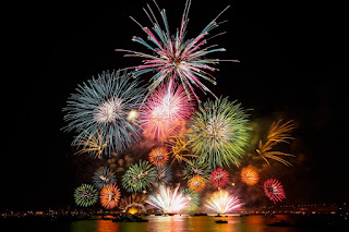 Foto. No céu escuro, sobre a cidade de Hiroshima, à beira do rio Ota, uma explosão de fogos de artifício em forma de um gigantesco arranjo floral colorido e rutilante. Na parte superior, as flores são maiores, no meio, menores e na base dois tufos em abertura de luzes brancas. Sobre a superfície do rio, algumas embarcações e o reflexo dos fogos.  Detalhe: “Hanabi” o termo vem das palavras japonesas Hana:“flor” e Bi: “fogo” combinadas. Os fogos de artifício exibidos no Japão são famosos por sua perfeição artística.  Desejamos um 2022 repleto de Hanabi a todos!