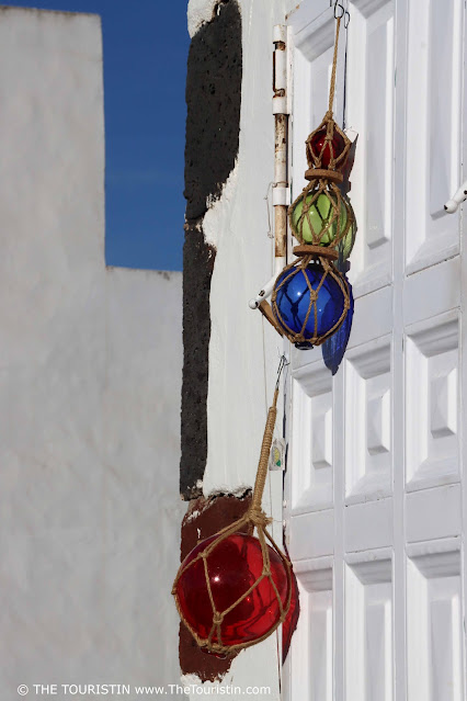 Large red, blue, and green glass baubles decorated in a row in a beige fishing net, hanging on the white door of a white cottage that is built with large grey boulders on its corners.