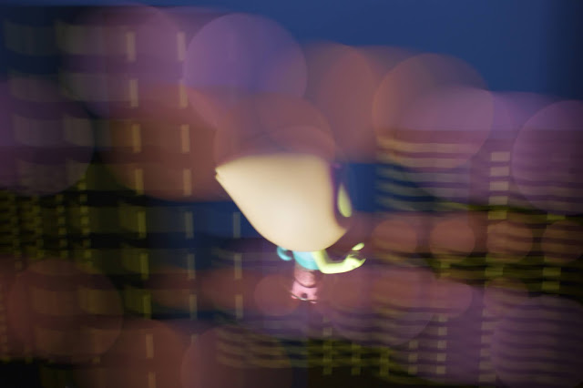 The background of this photo is blurred, but it shows several black city buildings with yellow windows against a dark blue night sky. Pale orange and pink lens flares are superimposed across the image. In the center of the image, a figurine of the character Pearl from Steven Universe (with light pink hair in a cone shape with the point in back, and wearing a light blue leotard) looks out over the cityscape and is not blurred.