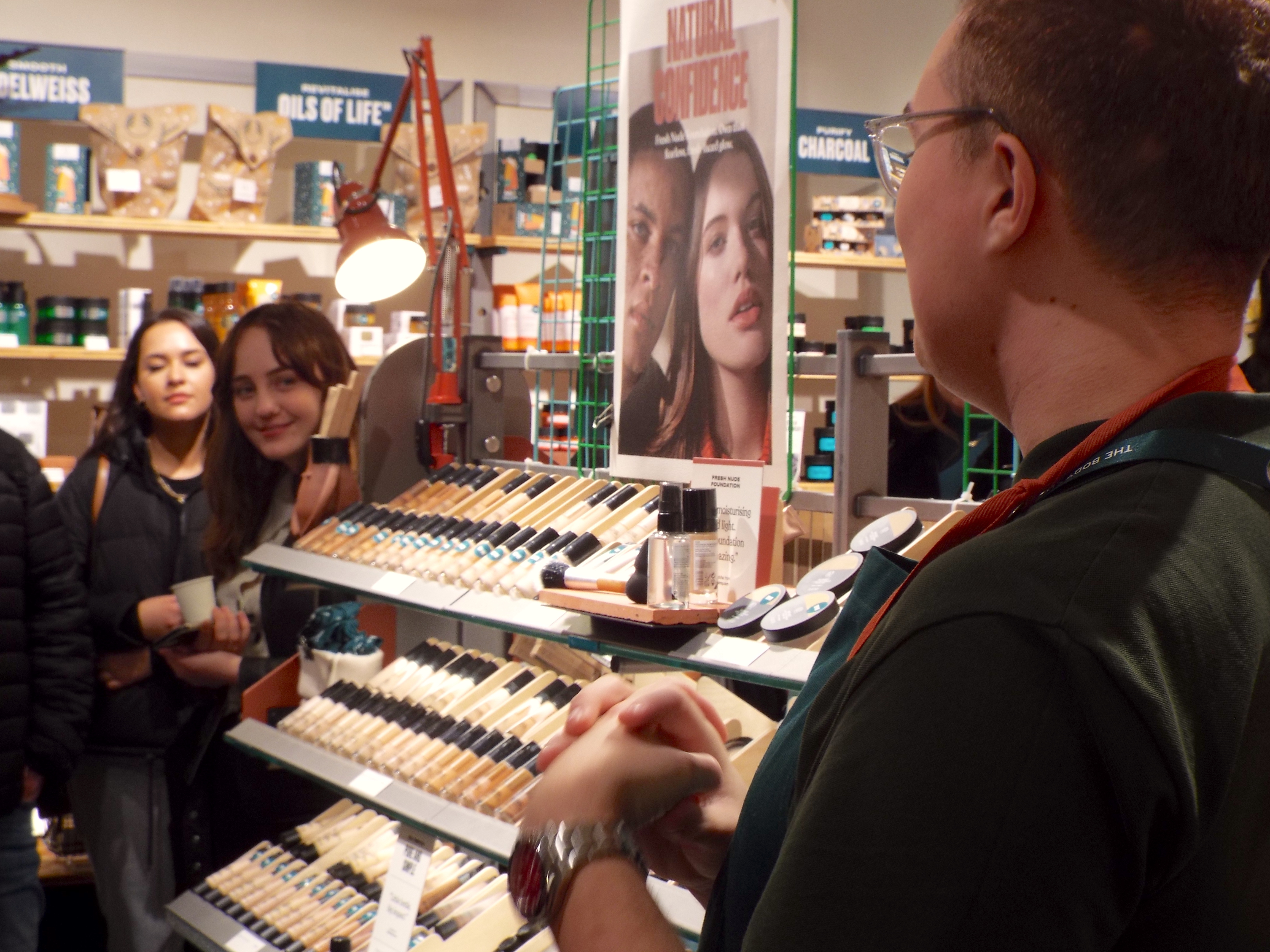 An array of foundations are on display at the new makeup counter, with Ryan stood at the top discussing the new makeup releases and a girl eagerly listens, poking her head around the corner intriguingly.