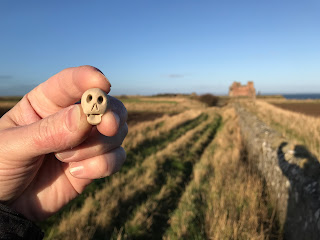 A photo of a small ceramic skull (Skulferatu 64) being held up with a wall, a field and Tantallon Castle in the background.  Photo by Kevin Nosferatu for the Skulferatu Project.